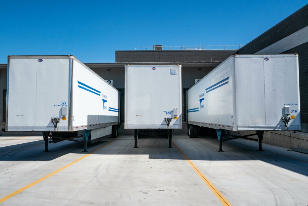 Three large white freight trailers parked at a loading dock, lined up side by side, with bright blue skies in the background. The trailers are marked with blue stripes and identification numbers, positioned for loading or unloading at a modern industrial facility